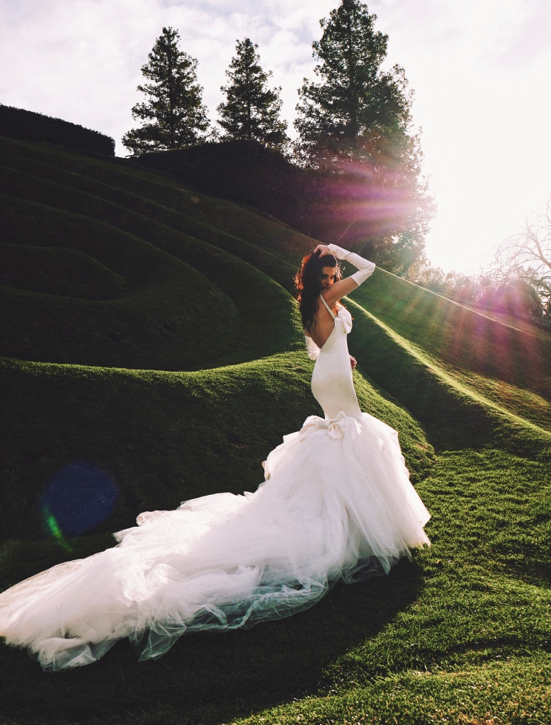A model wears a satin mermaid wedding dress with bow and detachable tulle skirt in a grass labyrinth.